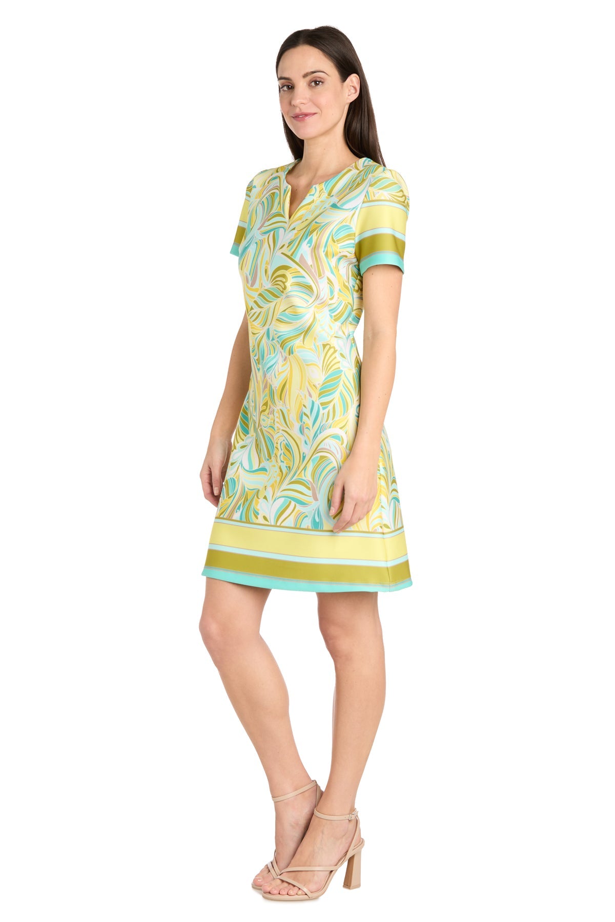 A woman wears the R&M Richards 4026 Short Yellow Print Party Dress, a knee-length, short-sleeve cocktail dress with an abstract pattern, paired with nude heeled sandals.